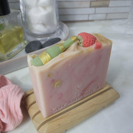  A bar of soap stands upright on a wooden soap dish on a bathroom counter with accessories.  The soap has a pale pink base colour with dark pink and gold swirls.  The top of the soap has embellishments of a green champagne bottle with gold detail along with half a strawberry.