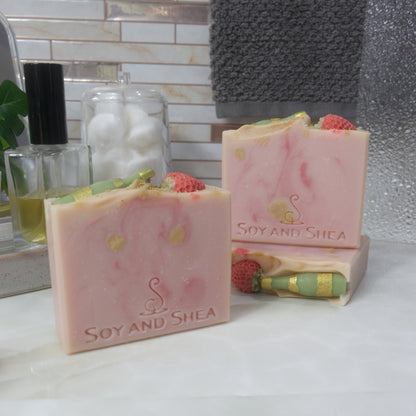 3 bars of soap are presented on a bathroom countertop next to a bottle of perfume and jar of cotton balls.   One soap stands upright to the front left and behind to the right, another soap lays flat showing the top of the soap, with a third standing on it.  The soaps are a pale pink, with dark pink and gold swirls.  The soaps feature a green bottle and strawberry on their tops made with soap