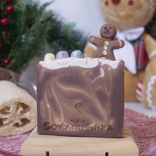 The Sugar & Spice Soap Bar (Christmas Edition) by Soy and Shea is a festive handmade soap featuring swirled brown and cream colors. It showcases a small gingerbread figure adorned with colorful candy-like decorations on top, set against a Christmas-themed background with greenery and a gingerbread decoration—ideal for enthusiasts of plant-based soaps.