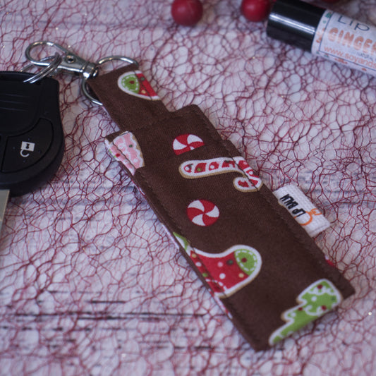 A lip balm holder lies on a white surface covered in red mesh and is attached to a car key with a "lobster claw" clip.  To the side is a tube of lip balm surrounded by christmas greenery and red berries.  The Clip Balm is a rich brown with images of candy cane, stockings and Christmas tress in traditional christmas colours