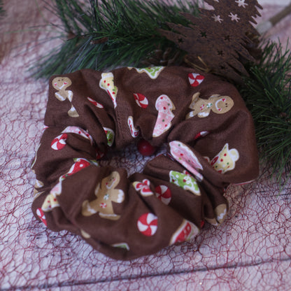 A colorful scrunchie sits on white surface covered with red mesh and rests against a pine branch. The scrunchie features an array of christmas themed sugar cookie images including Gingerbread men and trees along with peppermint swirl candies all on a rich brown background