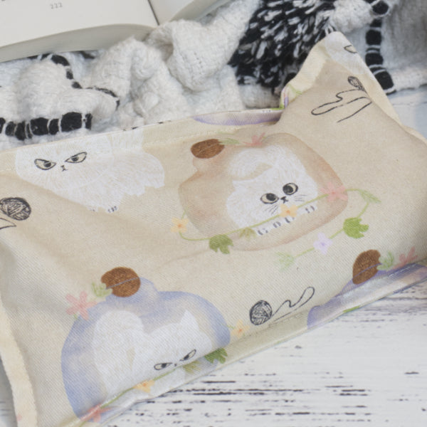The Sulky Kittens Mini Heat Pack by Soy and Shea is a beige fabric pouch with fluffy white cats in pastel vases and yarn balls—a cute, cozy accessory for warmth, shown on a white wooden surface beside a knit blanket and open book.