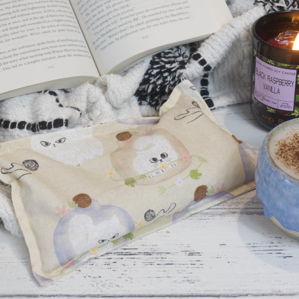 A cozy scene with an open book, Sulky Kittens Mini Heat Pack by Soy and Shea, a lit "Black Raspberry Vanilla" candle, and a frothy cup of coffee on a white wooden surface.  The small rectangular Heat Pack is beige with small grumpy looking kittens trapped under glass domes or playing with b alls of wall