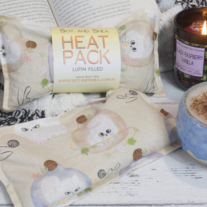 The Soy and Shea Sulky Kittens Mini Heat Pack with a grumpy looking kittens stuck under a glass bowl with balls of wool,  rests on a white surface next to a soft black-and-white blanket, a lit candle, and a ceramic mug.