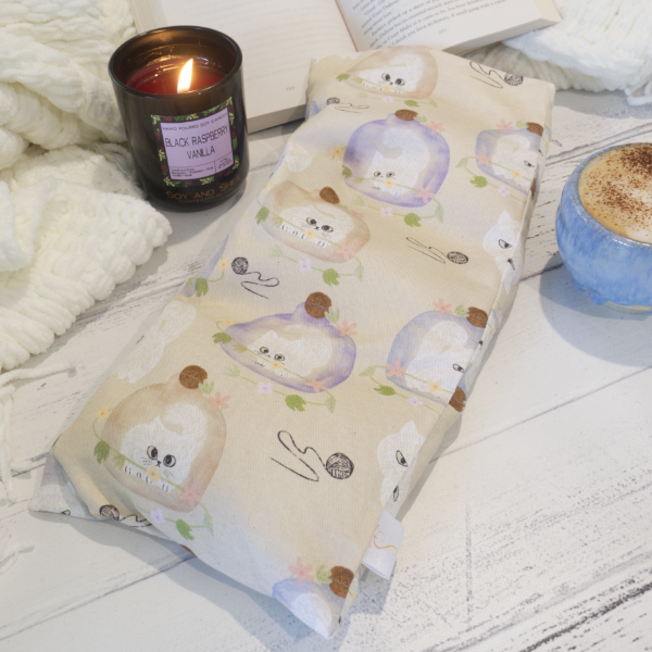 A rectangular heat pack with a beige background featuring illustrations of white kittens trapped in purple or brown vases surrounded by leaves and balls of wool. The pack rests on a wooden surface with a burning candle and a cup of coffee with an open book in the background, suggesting a cosy atmosphere.