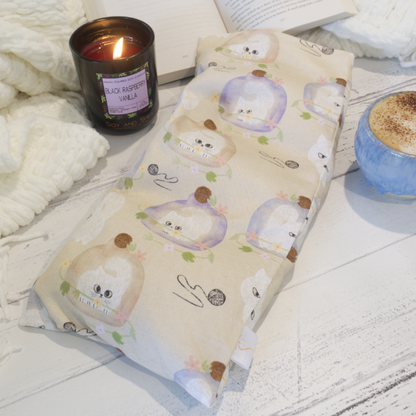 A rectangular heat pack with a beige background featuring illustrations of white kittens trapped in purple or brown vases surrounded by leaves and balls of wool. The pack rests on a wooden surface with a burning candle and a cup of coffee with an open book in the background, suggesting a cosy atmosphere.