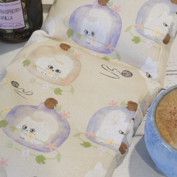 A close up of the Sulky Kitten Heat pack focusing on the fabric design.  Small grumpy looking white kittens are in a glass vases of either purple of brown surrounded by balls of wool all on a beige background