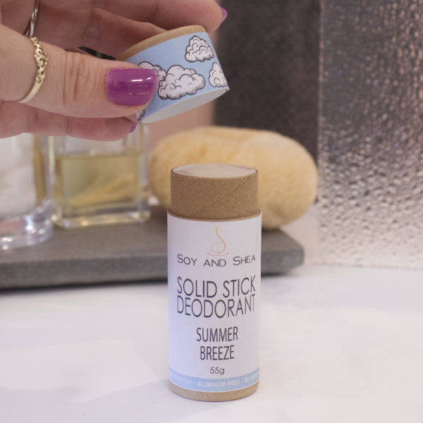 A cardboard tube labelled SOLID STICK DEODORANT stands upright on a bathroom counter top with a blurred background.  A hand hovers above the tube holding the lid in the finger tips revealing  a cream balm inside