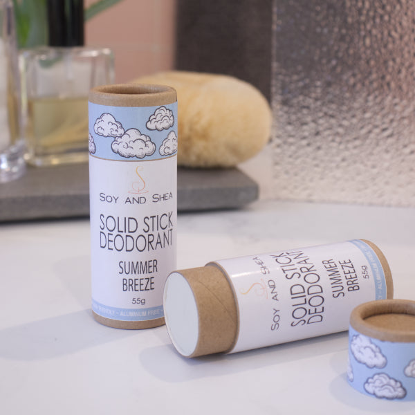 Two cardboard tubes labelled with SOLID STICK DEODORANT rest on a bathroom counter top with a blurred background.  One tube stands upright on the bench while another lays flat with the lid removed to reveal an off-white balm inside.  The label on the lid is light blue with white clouds.