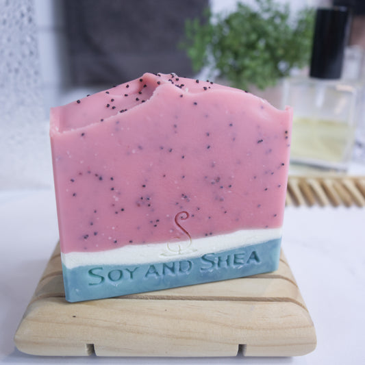 A soap bar sits on a wooden soap dish with a blurred bathroom background.  The soap has a dark green base layer with a narrow white layer followed by a large red layer with black speckles to look like a slice of watermelon.