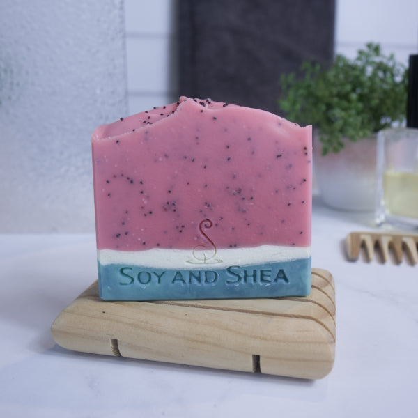 A bar of soap designed to look like a slice of watermelon sits on a wooden soap dish on a bathroom counter top.  The lower quarter of the soap is layered with a dark green base and white top.  The top three quarters is red with black speckles from poppy seeds.