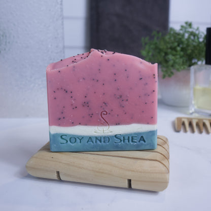 A bar of soap designed to look like a slice of watermelon sits on a wooden soap dish on a bathroom counter top.  The lower quarter of the soap is layered with a dark green base and white top.  The top three quarters is red with black speckles from poppy seeds.