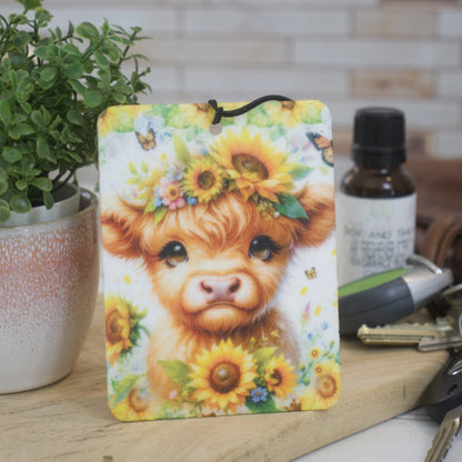 A car air freshener featuring an illustration of a Highland cow with sunflowers and other flowers around its head.  In the background is a bottle of scent and a set of keys