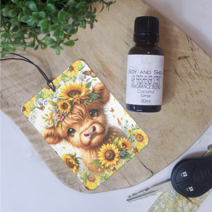 A car air freshener featuring an illustration of a Highland cow with sunflowers and other flowers around its headlays on a wooden board next to a bottle of Air Freshener scent.  