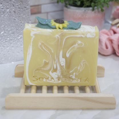 A yellow soap bar with white swirls sits on a wooden soap dish on a bathroom counter.  The top of the soap features sunflower embellishment surrounded by leaves