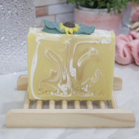 A yellow soap bar with white swirls sits on a wooden soap dish on a bathroom counter.  The top of the soap features sunflower embellishment surrounded by leaves