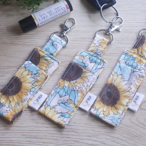 A selection of three clip balms showing variations in fabric design, lie on a wooden side with the middle Clip Balm attached to a car key.  A lip balm lays near-by along with some greenery.  The Clip balms have a large sunflower print in pastel shades on a cream background.  Each Lip Balm holder shows a slight variation in pattern design