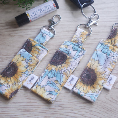 A selection of three clip balms showing variations in fabric design, lie on a wooden side with the middle Clip Balm attached to a car key.  A lip balm lays near-by along with some greenery.  The Clip balms have a large sunflower print in pastel shades on a cream background.  Each Lip Balm holder shows a slight variation in pattern design