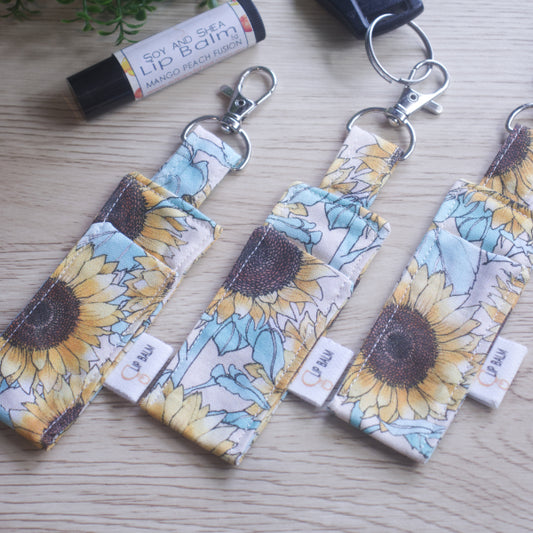 A selection of three clip balms showing variations in fabric design, lie on a wooden side with the middle Clip Balm attached to a car key.  A lip balm lays near-by along with some greenery.  The Clip balms have a large sunflower print in pastel shades on a cream background.  Each Lip Balm holder shows a slight variation in pattern design