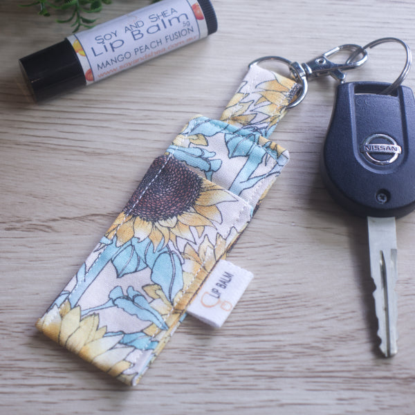A lip balm holder lies on a wooden surface attached to a car key with a "lobster claw" clip.  To the side is a tube of lip balm. The Clip Balm has a large sunflower print in pastel colours against a cream background