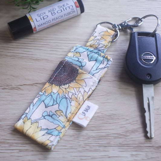 A lip balm holder lies on a wooden surface attached to a car key with a "lobster claw" clip.  To the side is a tube of lip balm. The Clip Balm has a large sunflower print in pastel colours against a cream background
