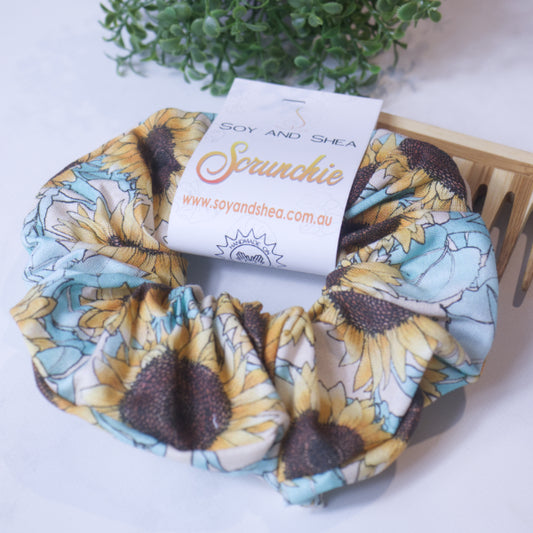 A colorful scrunchie sits on wooden surface resting against a wooden comb with greenery in the background.  A cardboard tag reads SOY AND SHEA SCRUNCHIE.    The scrunchie has a large sunflower print with pastel green leaves against a cream background
