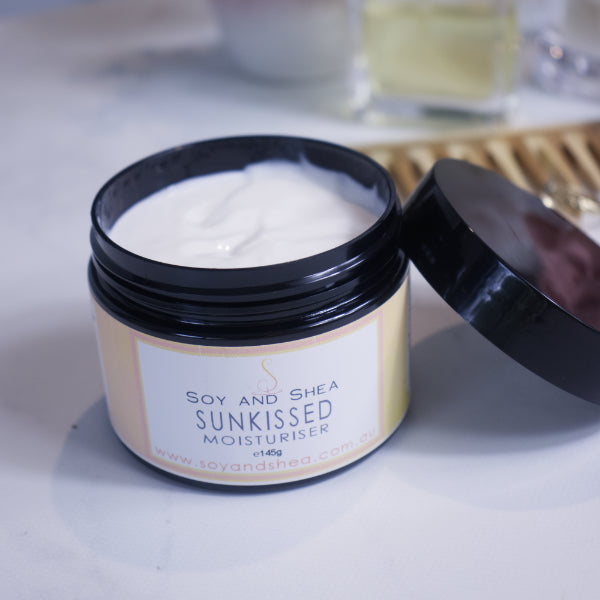 A black jar with yellow label reading SUNKISSED MOISTURISER sits on a white counter with a blurred background.  The black lid of the jar rests against the side of the jar revealing a white cream