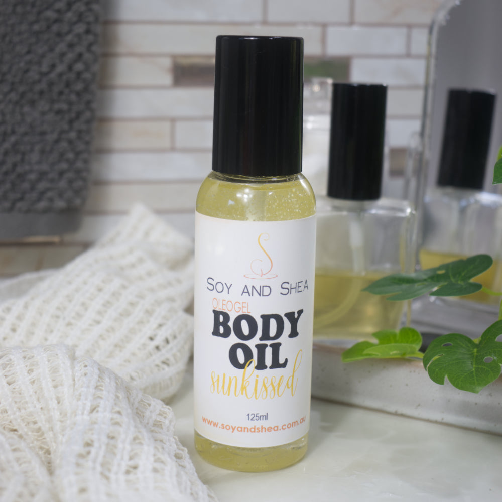 A clear bottle with black lid stands on a bathroom counter with a plant in the background.  The bottle is filled with a clear, golden gel and features a product label identifying it as Body Oil in Sunkissed Scent