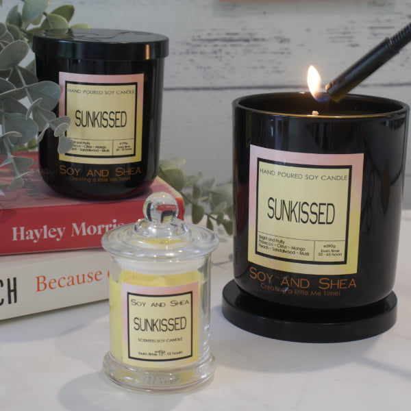 Three Soy and Shea branded candles all with labels reading 'Sunkissed'sit on a wooden surface.  A small candle with clear glass and yellow wax sits in front of a stack books, with a medium sized black jar sitting upon them.  The third black jar is the largest and is resting on its lid while a light holds a flame towards the wick.