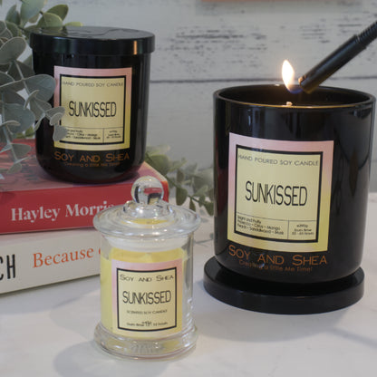 Three Soy and Shea branded candles all with labels reading 'Sunkissed'sit on a wooden surface.  A small candle with clear glass and yellow wax sits in front of a stack books, with a medium sized black jar sitting upon them.  The third black jar is the largest and is resting on its lid while a light holds a flame towards the wick.