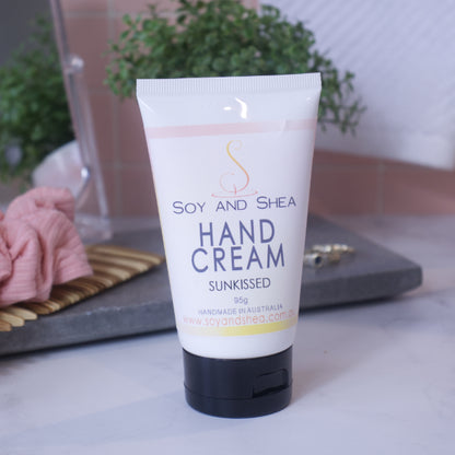 A tube of SOY AND SHEA HAND CREAM stands on a white surface against a pink tiled background, surrouned by potted plant, scrunchie and mirror.  The tube is white with a black flip top lid.