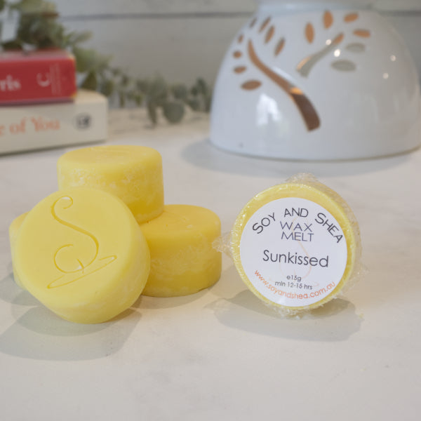 Three cylindrical yellow Melt Pucks are stacked up each other with the soy and shea logomark stamped on the tops.  To the right is a fourth melt puck that rests on its side with a label identifying it as Sunkissed.  Behind is a stack of books next to a lit oil warmer.