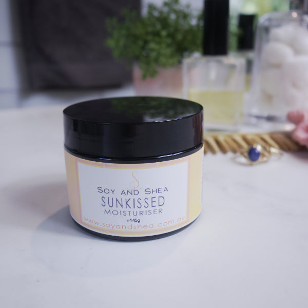 A black jar with yellow label reading SUNKISSED MOISTURISER sits on a white counter with a blurred background.  
