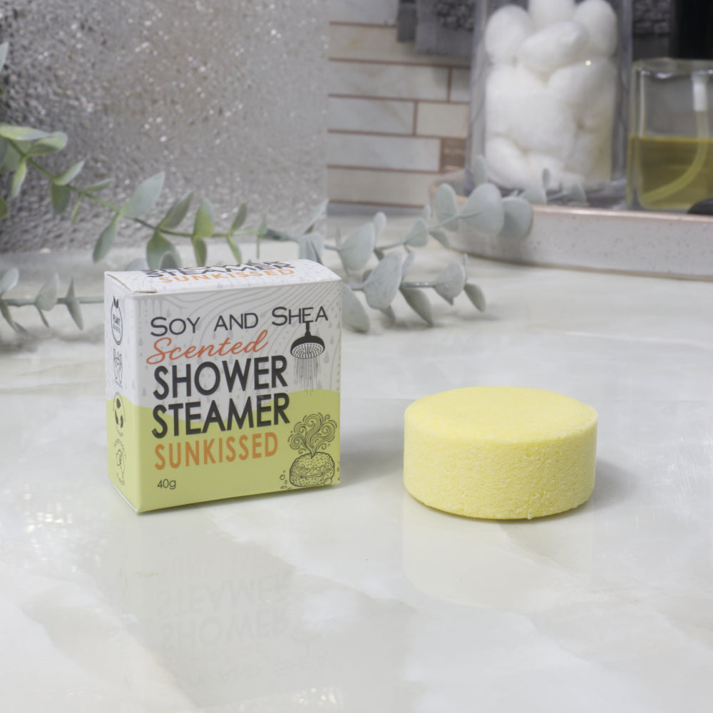 A boxed scented shower steamer and the steamer itself on a bathroom counter. The box reads 'Soy and Shea Scented Shower Steamer Sunkissed', and the round yellow steamer sits beside it.