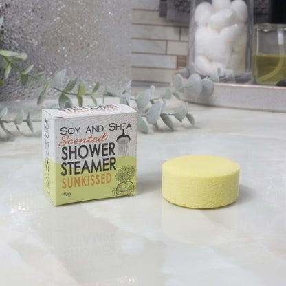 A boxed scented shower steamer and the steamer itself on a bathroom counter. The box reads 'Soy and Shea Scented Shower Steamer Sunkissed', and the round yellow steamer sits beside it.