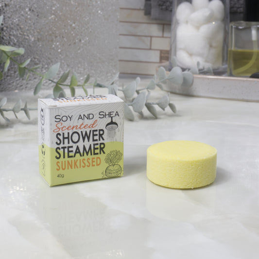 A boxed scented shower steamer and the steamer itself on a bathroom counter. The box reads 'Soy and Shea Scented Shower Steamer Sunkissed', and the round yellow steamer sits beside it.