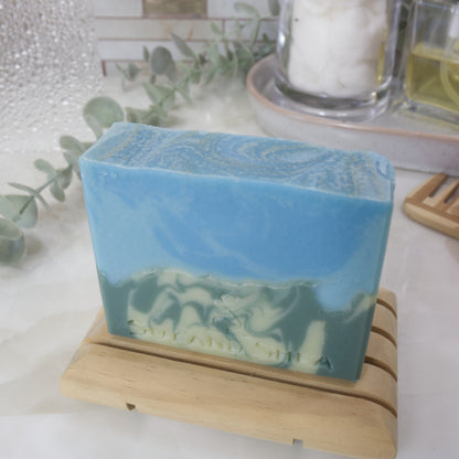 A bar of soap stands on a wooden soap rack on a bathroom counter top with a blurred background of greenery and bottles.  The soap has two layers with the bottom half having a swirl of light and dark green lookin glike a mountain side.  The top has a swirl of bright and light blue.  The top of the soap is flat with gold swirls through the blue