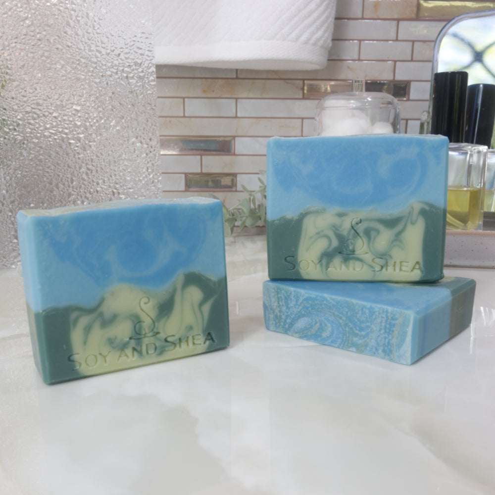 Three bars of soap are displayed on a bathroom counter top.  One soap  lies flat on the surface revealing a flat top soap that is blue with gold swirls.  A second soap stands on top of this bar with a third standing upright to the left.  The upright soaps show a design of two layers with the bottom layer having a swirl of light and dark green looking like a meadow on a mountain side, while the top layer is of light and bright blue swirls.