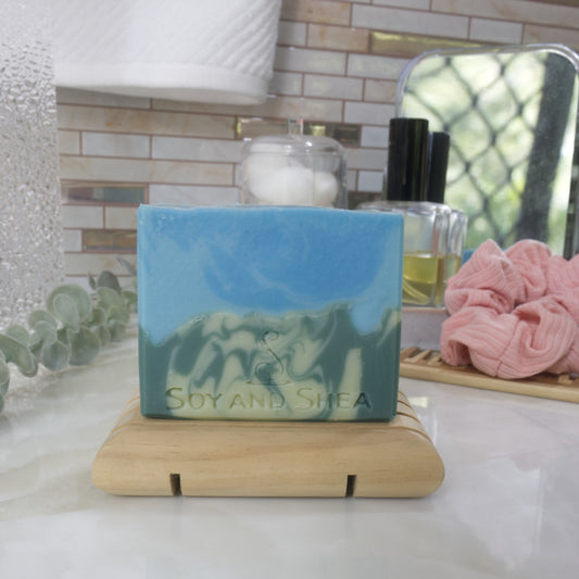 A bar of soap stands on a wooden soap rack on a bathroom counter top surrounded by miscellanous objects including scrunchie and bottle of perfume.  The soap has two layers with the bottom half having a swirl of light and dark green lookin glike a mountain side.  The top has a swirl of bright and light blue.
