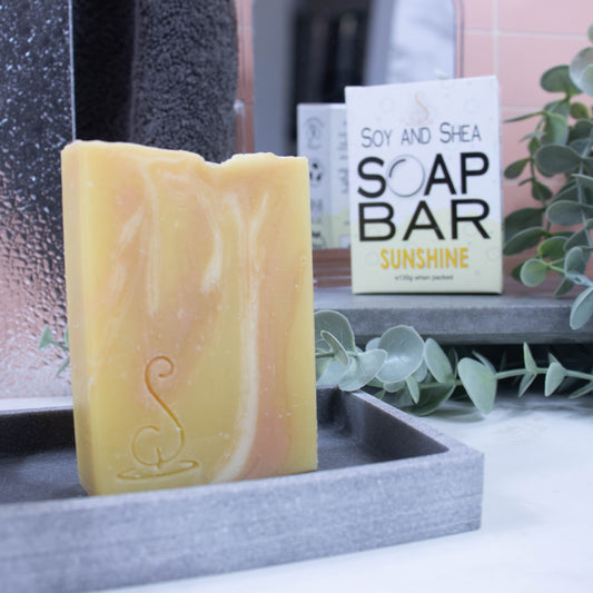 A tall rectangular bar of soap sits on a grey soap dish. The soap is yellow with gold and white streaks.  Behind the soap is a shower screen and towel along with a box the soap was presented in. The box has a white top with pale yellow bubble border.