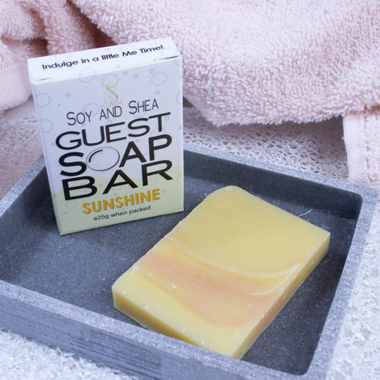 A grey stone soap dish sits next to a pink towel.  On the dish stands a white box with a border of yellow bubble foam across the bottom and text GUEST SOAP BAR.  Next to it lies a small piece of soap that is yellow with dark yellow and white wisps 