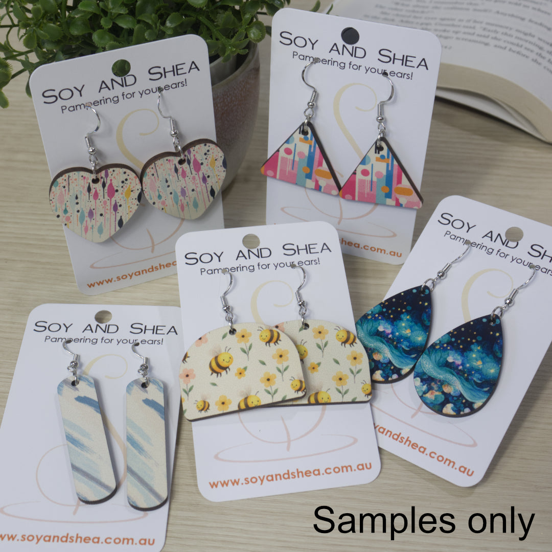 A collection of various earrings are displayed on their cardboard backing on a bench top.  The earrings are of various shapes and sizes with bright patterns.  All have silver hooks