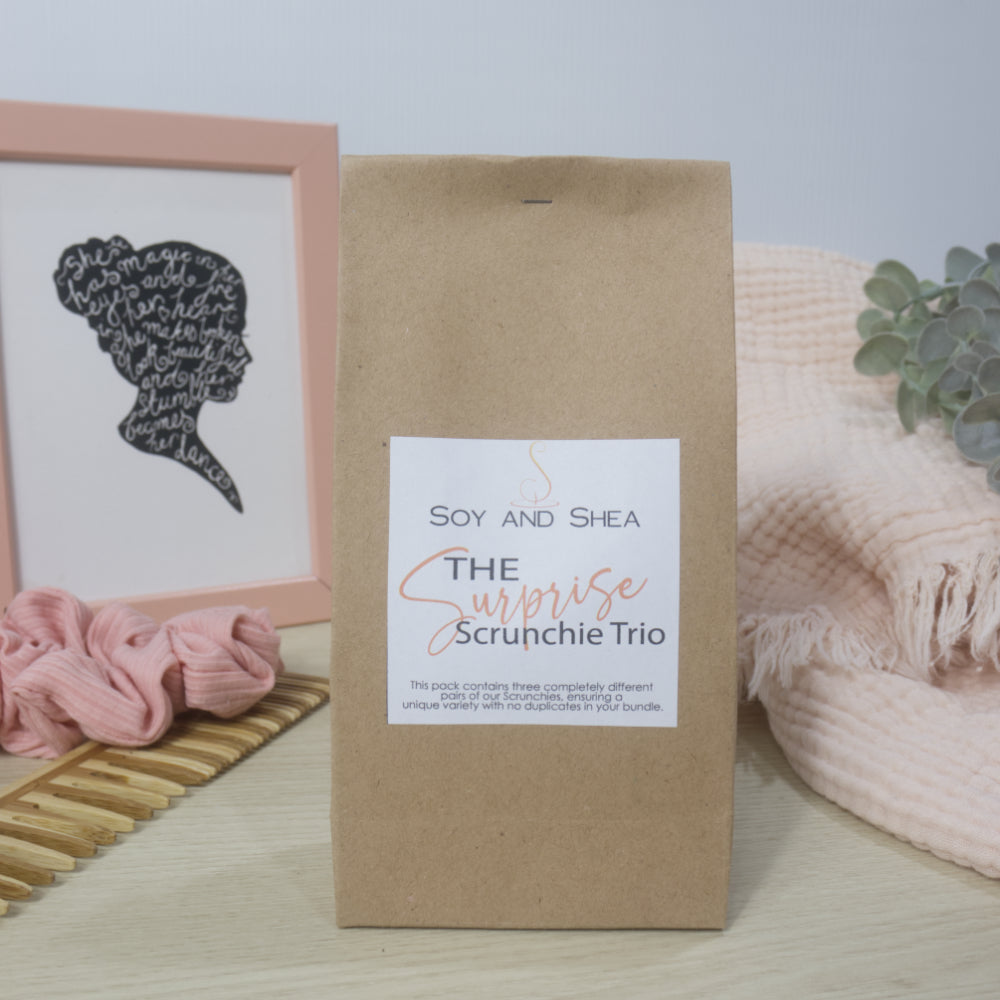 A brown paper bag stands on a bench labelled as The Surprise Scrunchie trio.  It is surrounded by a pink blanket, a photoframe and a comb with scrunchie