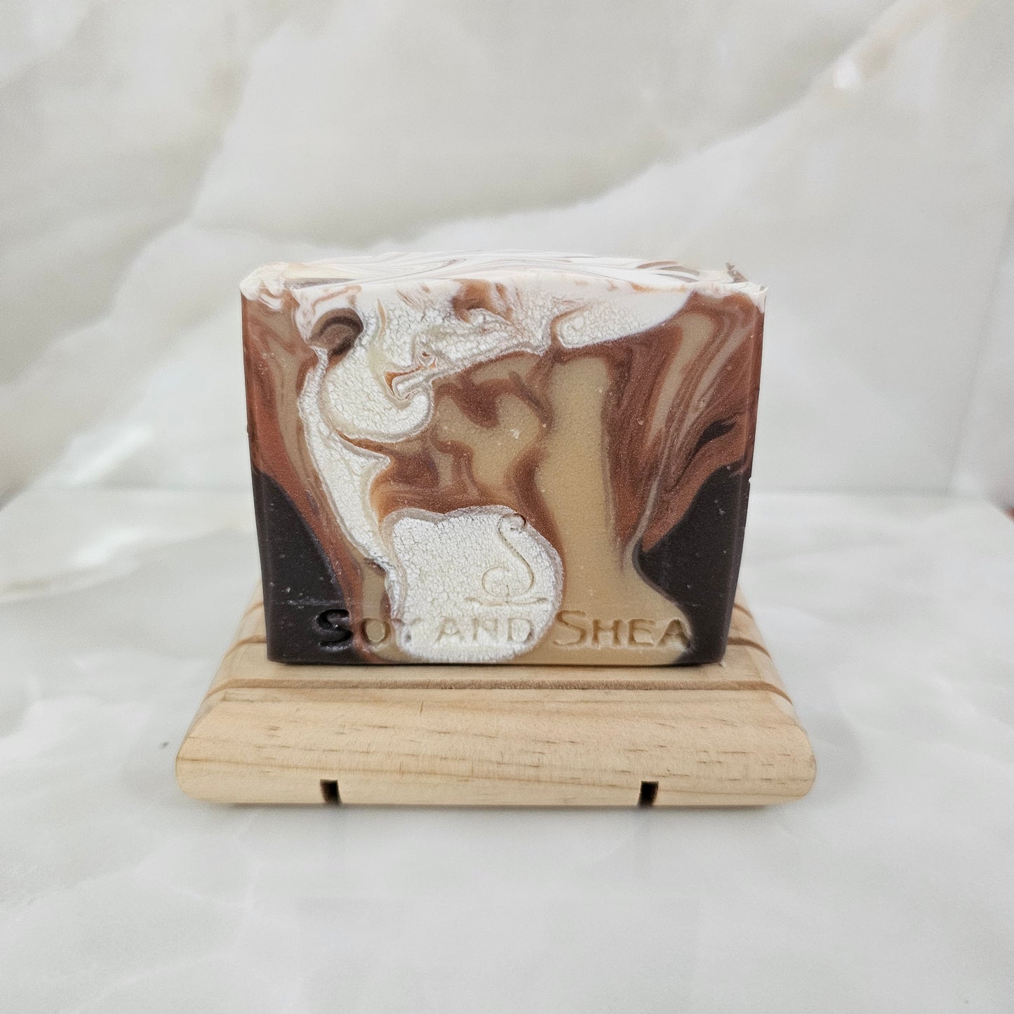 A soap bar with drops of colour including white and shades of brown, sits on wood stand with a marble white background