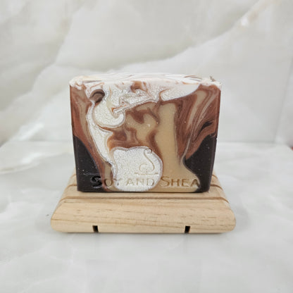 A soap bar with drops of colour including white and shades of brown, sits on wood stand with a marble white background