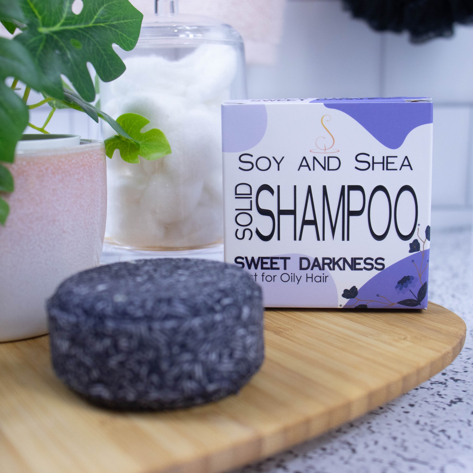On a wooden tray sits a round purple Shampoo bar and its cardboard box which is white with an abstract purple design.  Next to it is a potted plant and a jar of cotton balls against a white tile background.