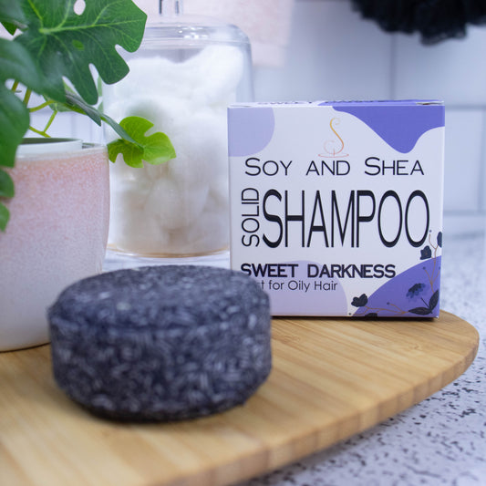 On a wooden tray sits a round purple Shampoo bar and its cardboard box which is white with an abstract purple design.  Next to it is a potted plant and a jar of cotton balls against a white tile background.