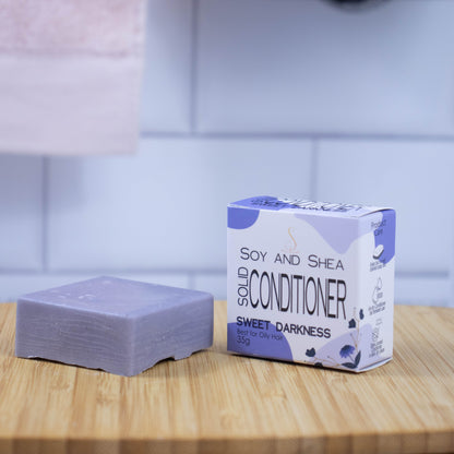 On a wooden tray sits a purple bar of conditioner next to a white box with a purple abstract pattern reading SOLID CONDITIONER against a blurred tiled background.