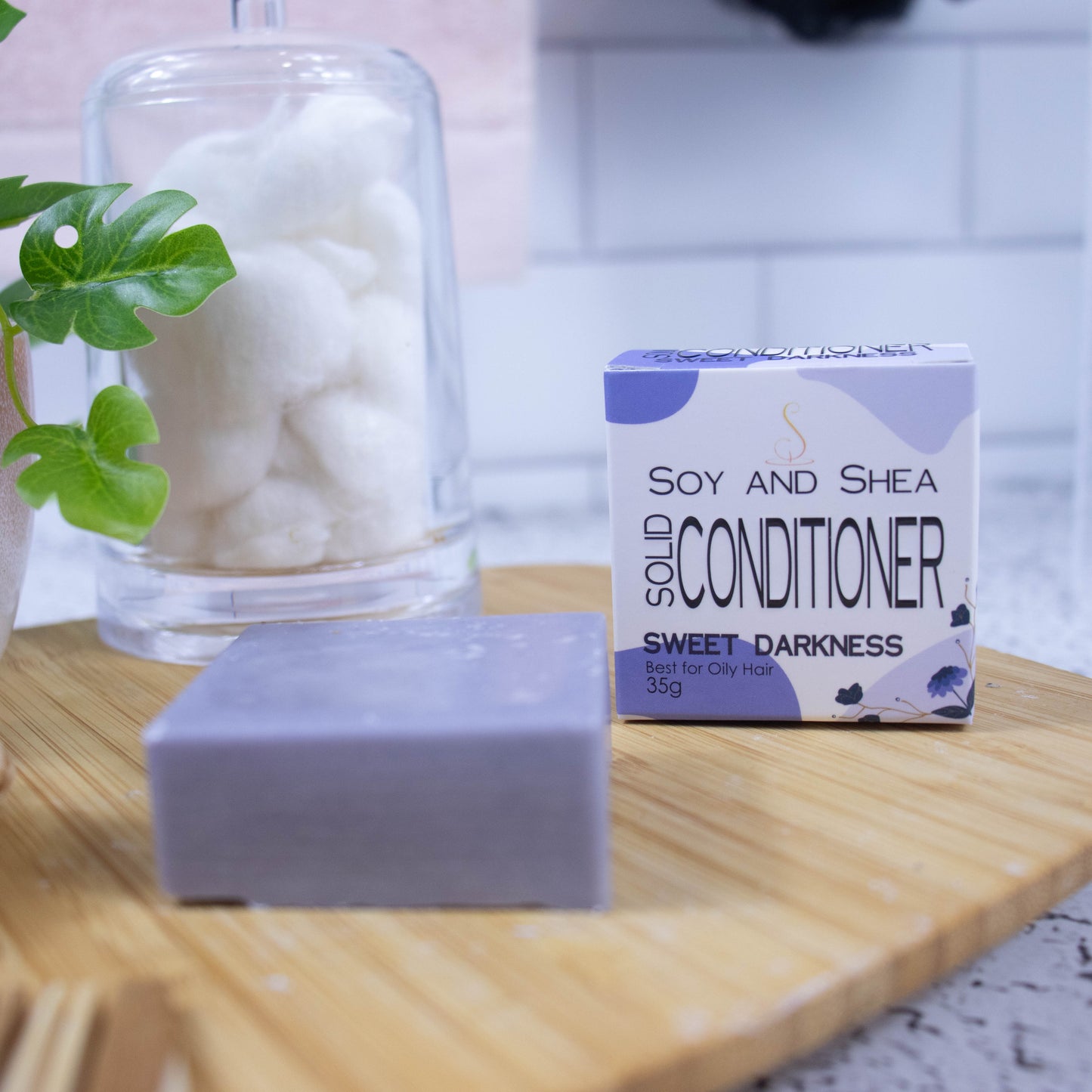 A wooden tray sits on a bathroom counter with a potted plant and jar of cotton balls against a blurred tiled background. Towards the front of the tray stands a white box with purple abstract pattern with text SOLID CONDITIONER and next to it stands a square purple bar of conditioner.