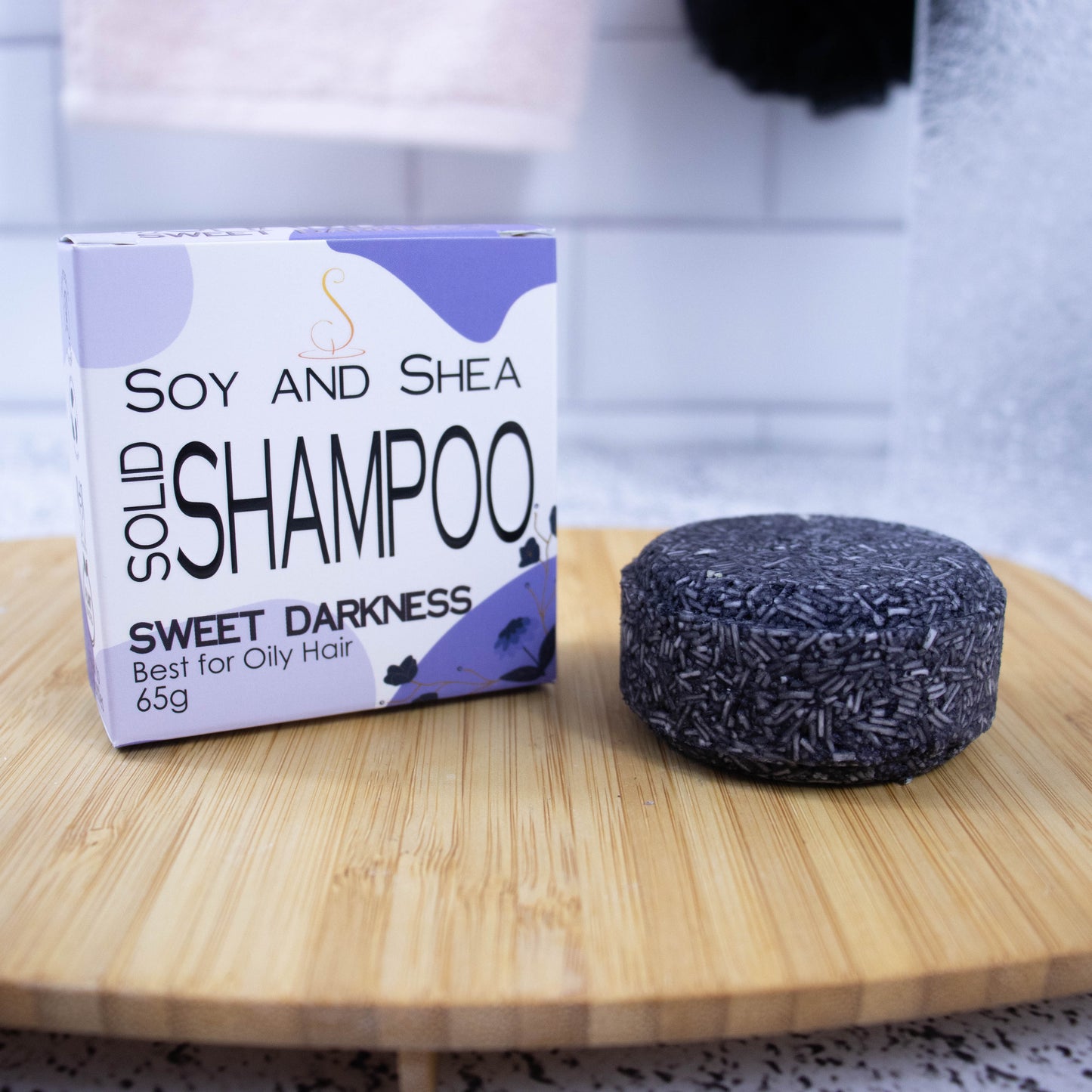 A white box with purple abstract pattern and the text SOLID SHAMPOO SWEET DARKNESS stands upright on a wooden tray with a blurred bathroom background.  Next to the box sits a round bar of solid shampoo.  The bar is dark purple with small white lines throughout.